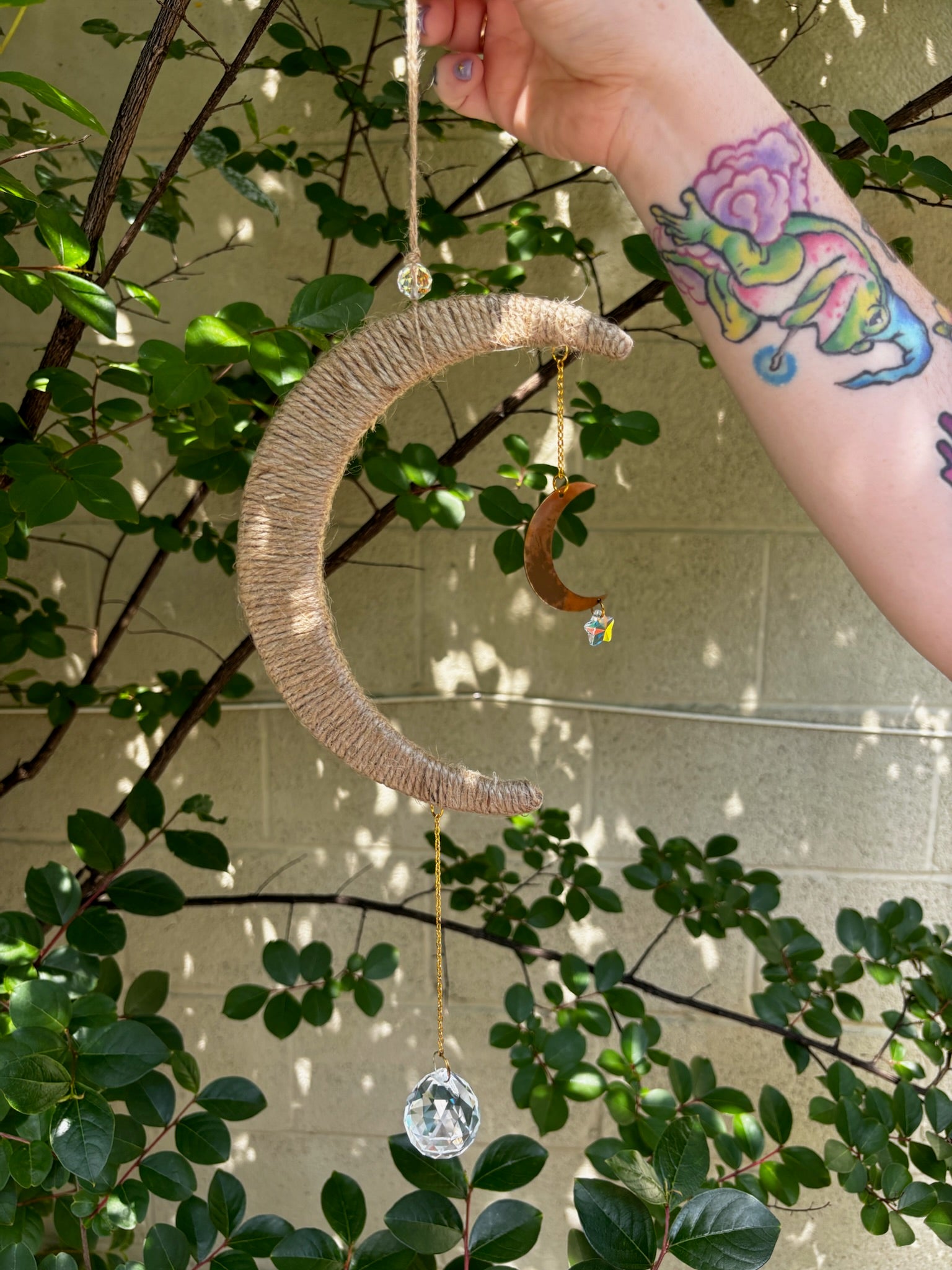 The Boho Woven Moon Suncatcher - Rustic Window Decor being held by someone in front of a cream-colored brick wall with greenery behind it.