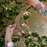 The Boho Woven Moon Suncatcher - Rustic Window Decor being held by someone in front of a cream-colored brick wall with greenery behind it.
