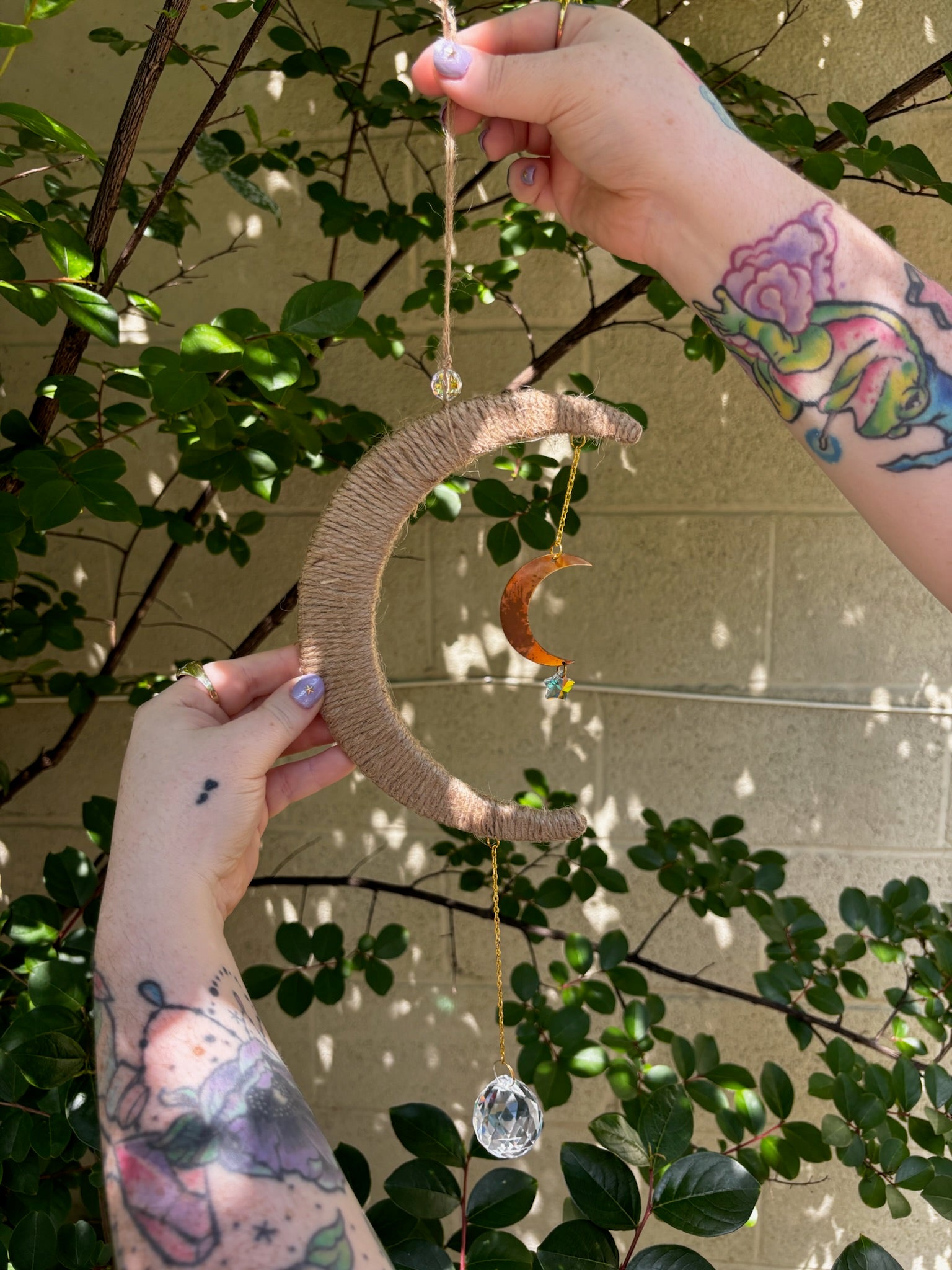 The Boho Woven Moon Suncatcher - Rustic Window Decor being held by someone in front of a cream-colored brick wall with greenery behind it.