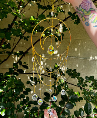Someone holding the Large Celestial Suncatcher in front of a cream-colored brick wall with greenery behind it.