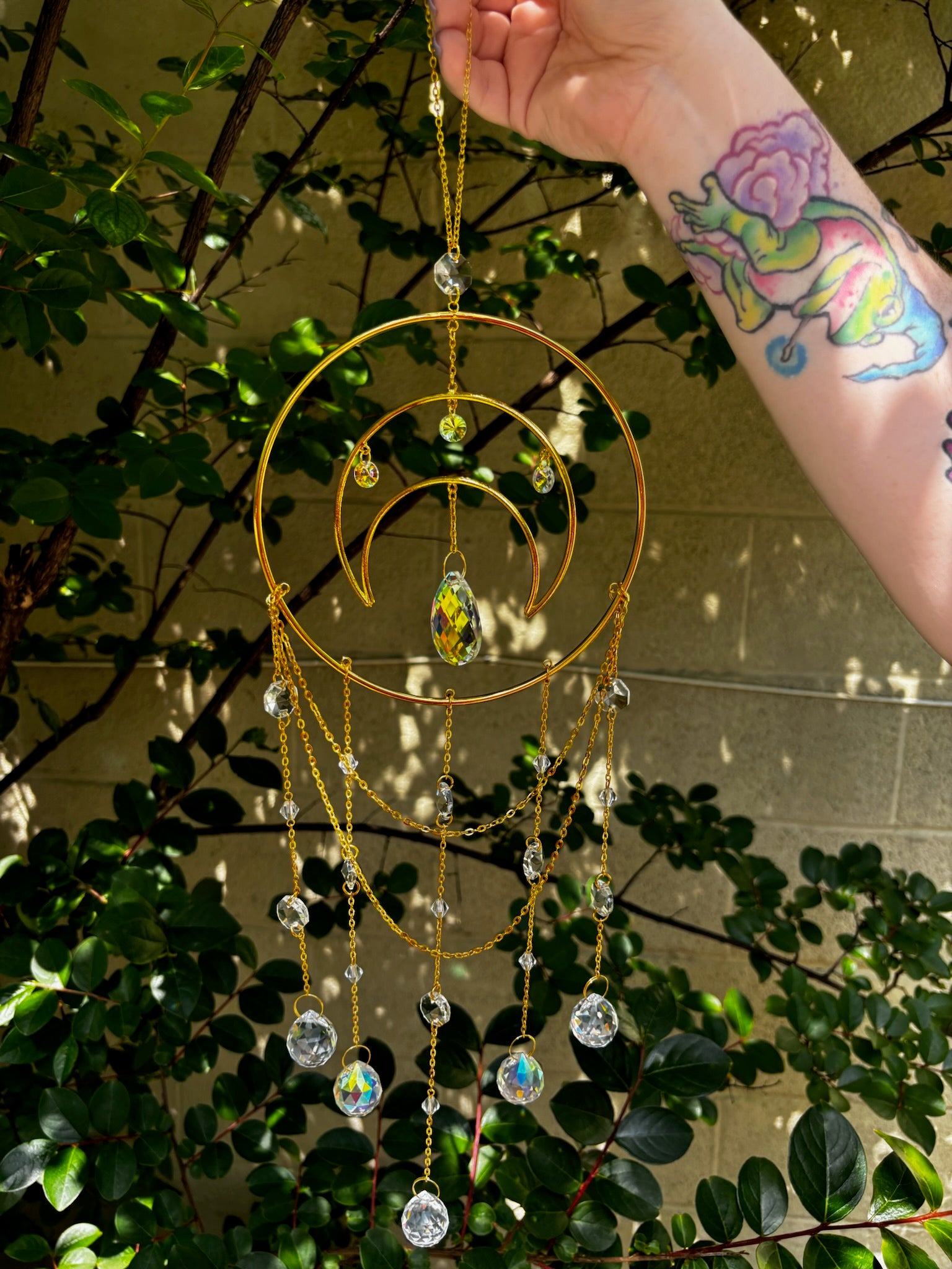 Someone holding the Large Celestial Suncatcher in front of a cream-colored brick wall with greenery behind it.