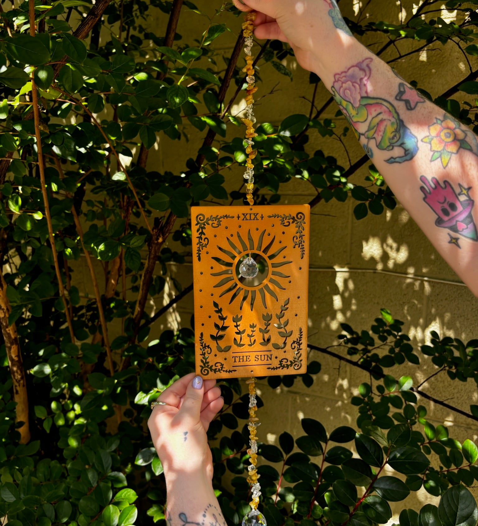 Someone holding the The Sun Wooden Tarot Suncatcher in front of a cream-colored brick wall with greenery.