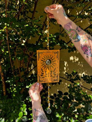 Someone holding the The Sun Wooden Tarot Suncatcher in front of a cream-colored brick wall with greenery.