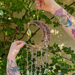 Someone holding the amethyst Moon Crystal Gemstone Suncatcher in front of a cream-colored brick wall with greenery.