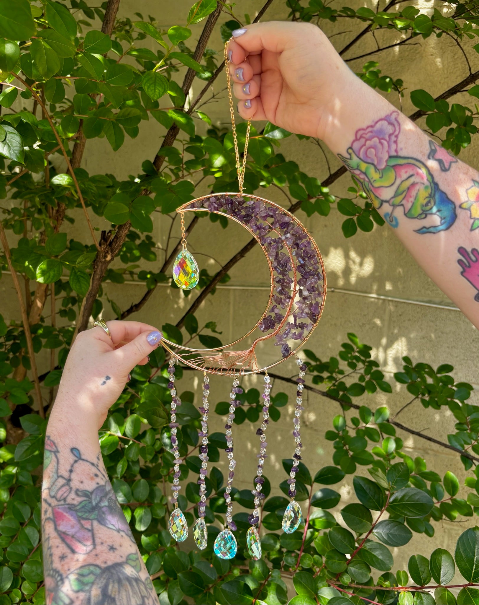 Someone holding the amethyst Moon Crystal Gemstone Suncatcher in front of a cream-colored brick wall with greenery.