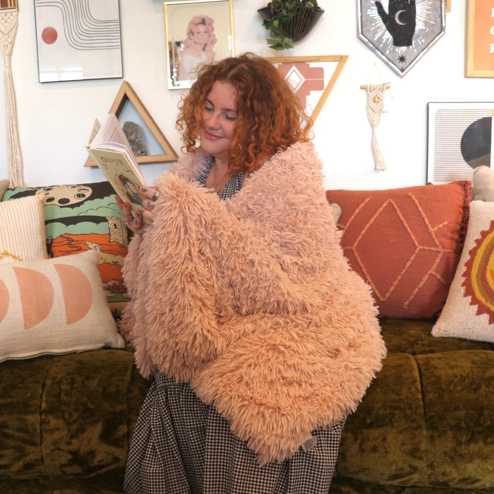 A woman reading on a couch and wrapped with the Pink Hero Dreamy Faux Fur Throw Blanket.