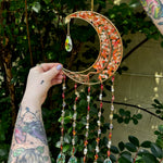 Someone holding the red agate Moon Crystal Gemstone Suncatcher in front of a cream-colored brick wall with greenery.
