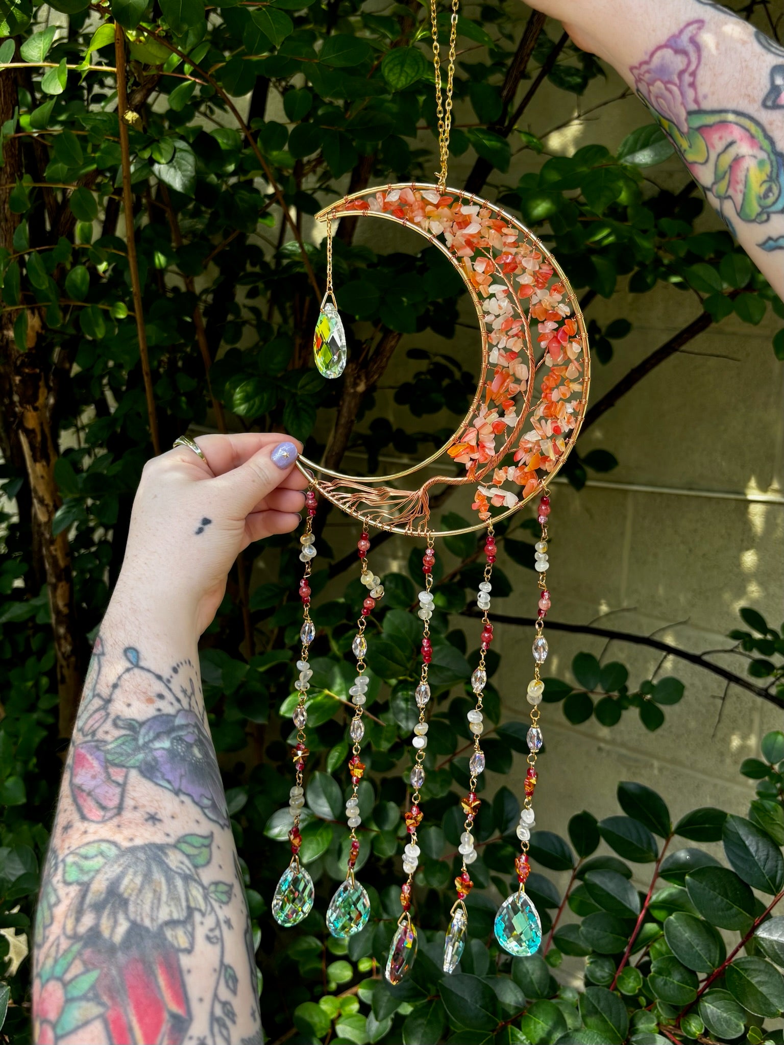 Someone holding the red agate Moon Crystal Gemstone Suncatcher in front of a cream-colored brick wall with greenery.