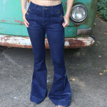 A woman wearing the Dark Denim Western Flared Jeans with Side Buttons with an orange, red, and brown crochet top all in front of a vintage Volkswagen bus.