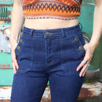 A woman wearing the Dark Denim Western Flared Jeans with Side Buttons with an orange, red, and brown crochet top all in front of a vintage Volkswagen bus.