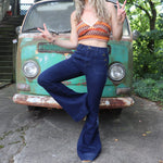 A woman wearing the Dark Denim Western Flared Jeans with Side Buttons with an orange, red, and brown crochet top all in front of a vintage Volkswagen bus.