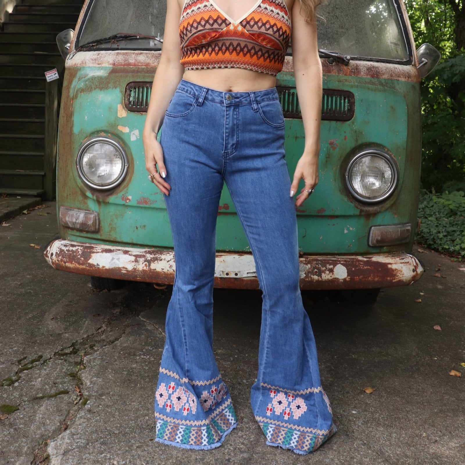 A woman wearing the Midtone Embroidered Denim Bell Bottoms and a red, orange, and brown crochet top in front of a vintage Volkswagen bus. 