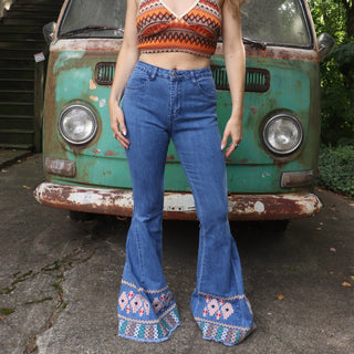 A woman wearing the Midtone Embroidered Denim Bell Bottoms and a red, orange, and brown crochet top in front of a vintage Volkswagen bus. 