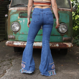A woman wearing the Midtone Embroidered Denim Bell Bottoms and a red, orange, and brown crochet top in front of a vintage Volkswagen bus. 