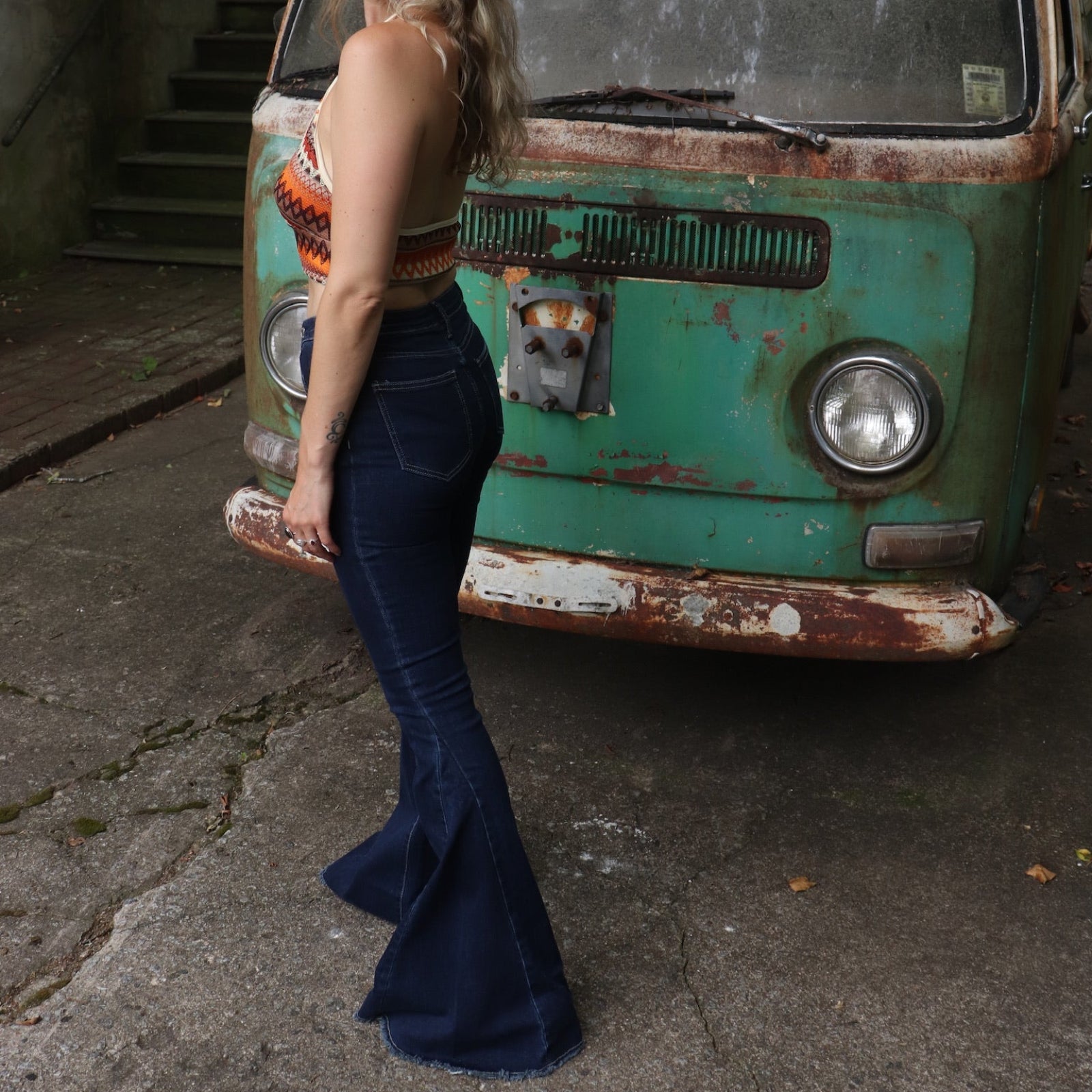 A woman wearing the Criss Cross Dark Wash Bell Bottom Jeans with an orange, red, and brown top all in front of a vintage Volkswagen bus.