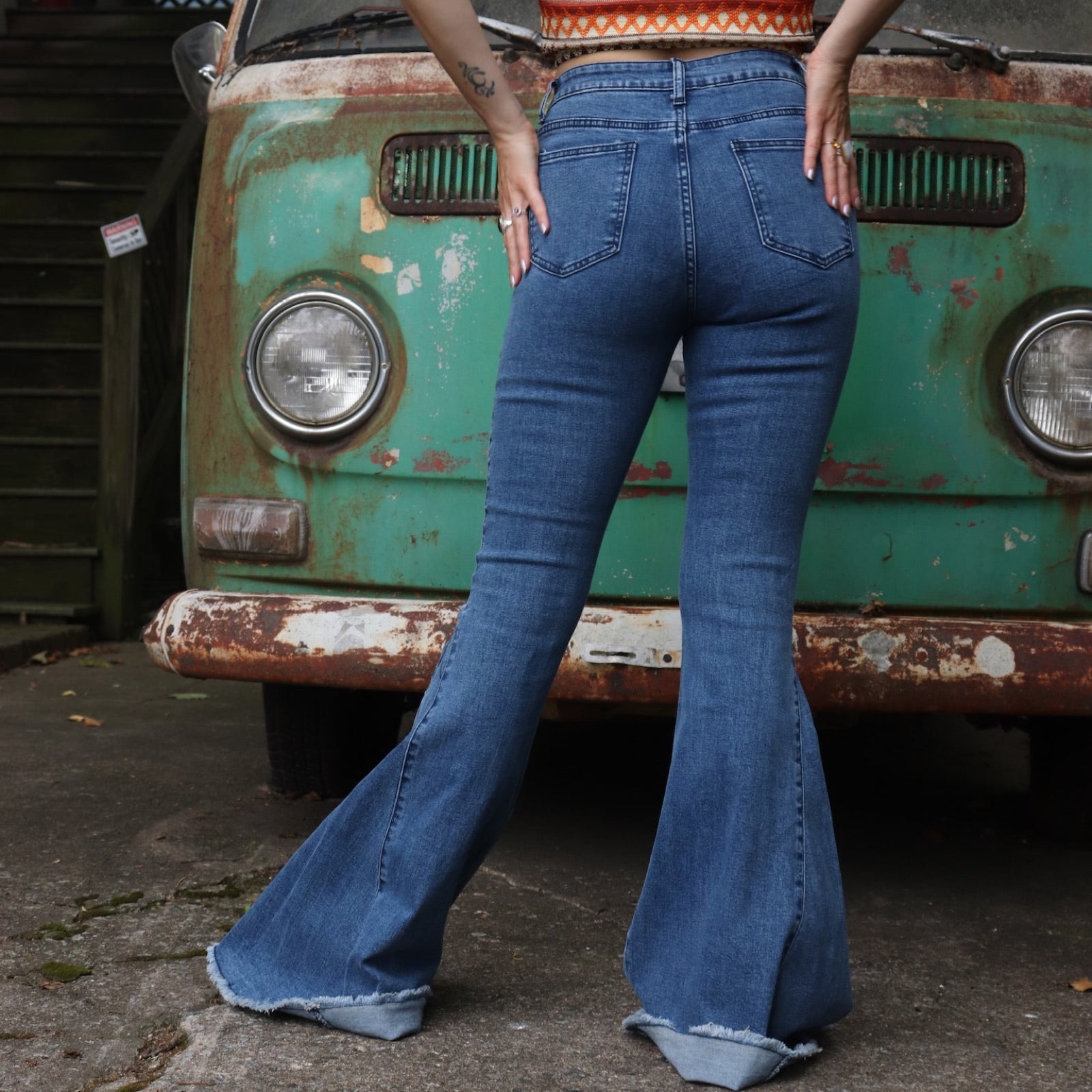 A woman wearing the Western Midtone Ripped and Fringed Stretchy Flared Jeans with a red, orange, and brown top all in front of a vintage Volkswagen bus.