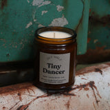 The Tiny Dancer candle in front of a white and teal rusty background.