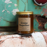 The Come Together candle in front of a white and teal rusty background.