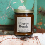 The Cherry Bomb candle in front of a white and teal rusty background.