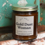 The Gold Dust Woman candle in front of a white and teal rusty background.