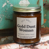 The Gold Dust Woman candle in front of a white and teal rusty background.