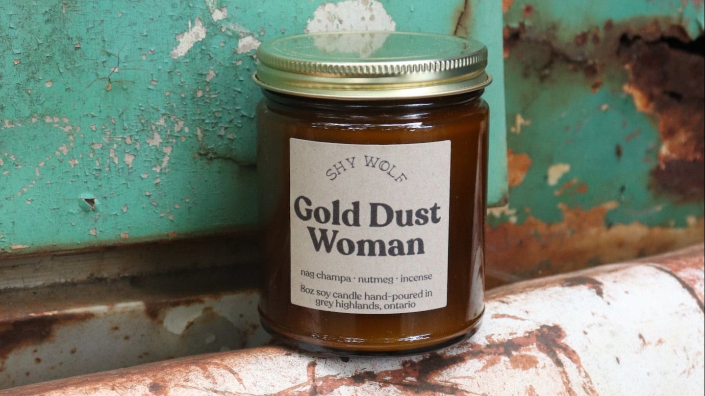 The Gold Dust Woman candle in front of a white and teal rusty background.