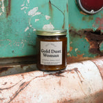 The Gold Dust Woman candle in front of a white and teal rusty background.