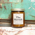 The Hermit candle in front of a white and teal rusty background.