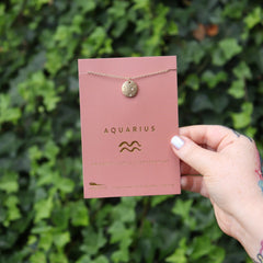 Someone holding the Aquarius 14k Gold Dipped Nickel-Free Zodiac Necklace on its card in front of greenery.