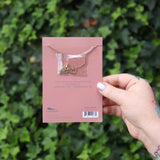 Someone holding the Aquarius 14k Gold Dipped Nickel-Free Zodiac Necklace on its card in front of greenery showing off the back.