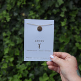 Someone holding the Aries 14k Gold Dipped Nickel-Free Zodiac Necklace on its card in front of greenery.