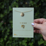 Someone holding the Virgo 14k Gold Dipped Nickel-Free Zodiac Necklace on its card in front of greenery.