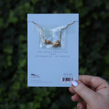 Someone holding the Libra 14k Gold Dipped Nickel-Free Zodiac Necklace on its card in front of greenery showing off the back.