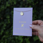 Someone holding the Pisces 14k Gold Dipped Nickel-Free Zodiac Necklace on its card in front of greenery.