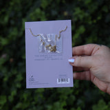Someone holding the Pisces 14k Gold Dipped Nickel-Free Zodiac Necklace on its card in front of greenery showing off the back.