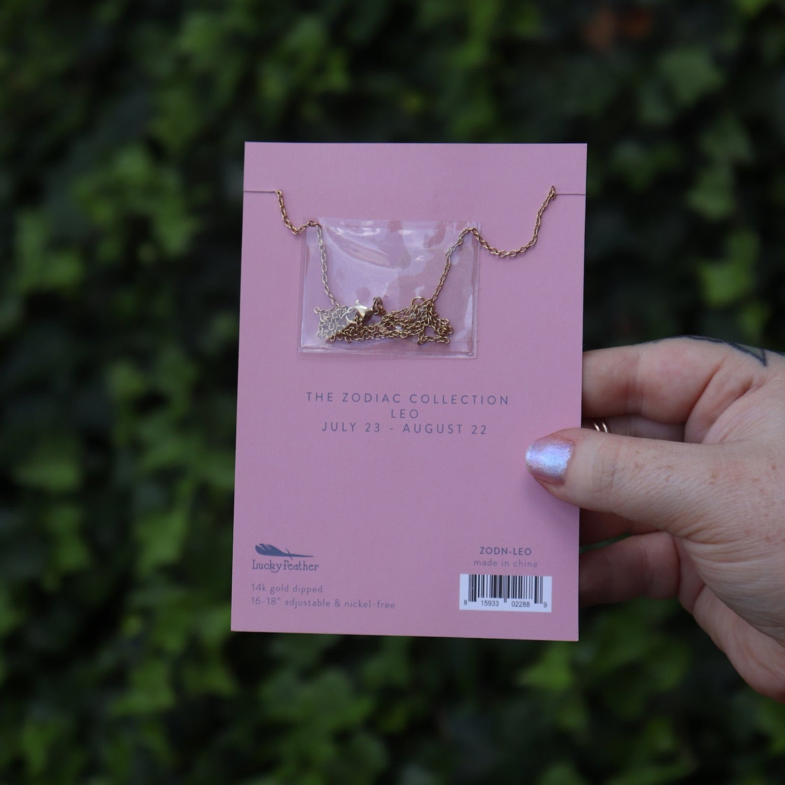 Someone holding the Leo 14k Gold Dipped Nickel-Free Zodiac Necklace on its card in front of greenery showing off the back.