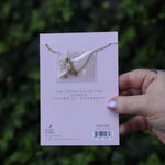 Someone holding the Scorpio 14k Gold Dipped Nickel-Free Zodiac Necklace on its card in front of greenery showing off the back.