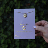 Someone holding the Scorpio 14k Gold Dipped Nickel-Free Zodiac Necklace on its card in front of greenery.