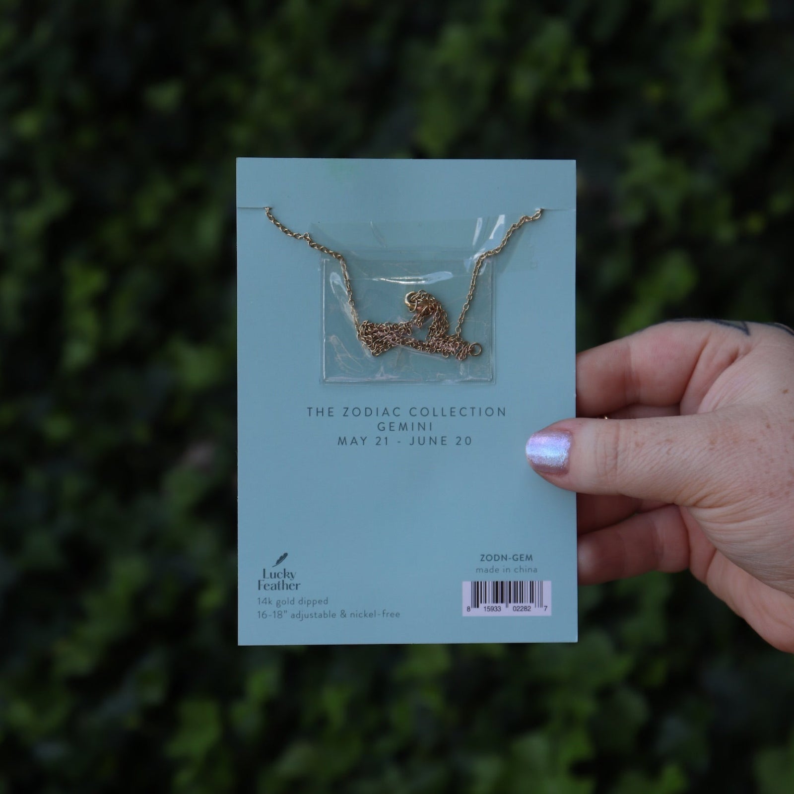 Someone holding the Aquarius 14k Gold Dipped Nickel-Free Zodiac Necklace on its card in front of greenery showing off the back.