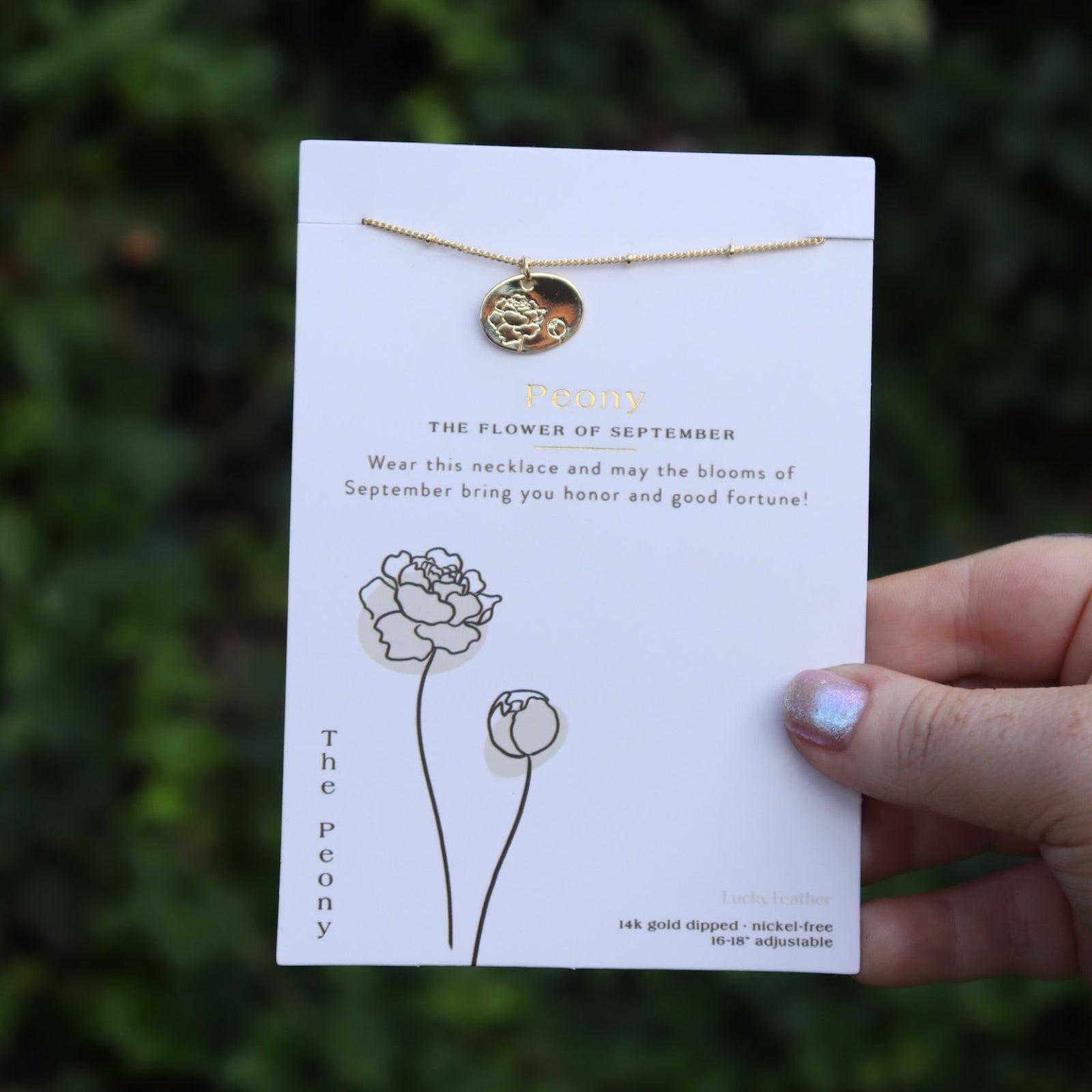 Someone holding the peony 14k Gold Dipped Nickel-Free Birth Month Flower Necklace in front of greenery.