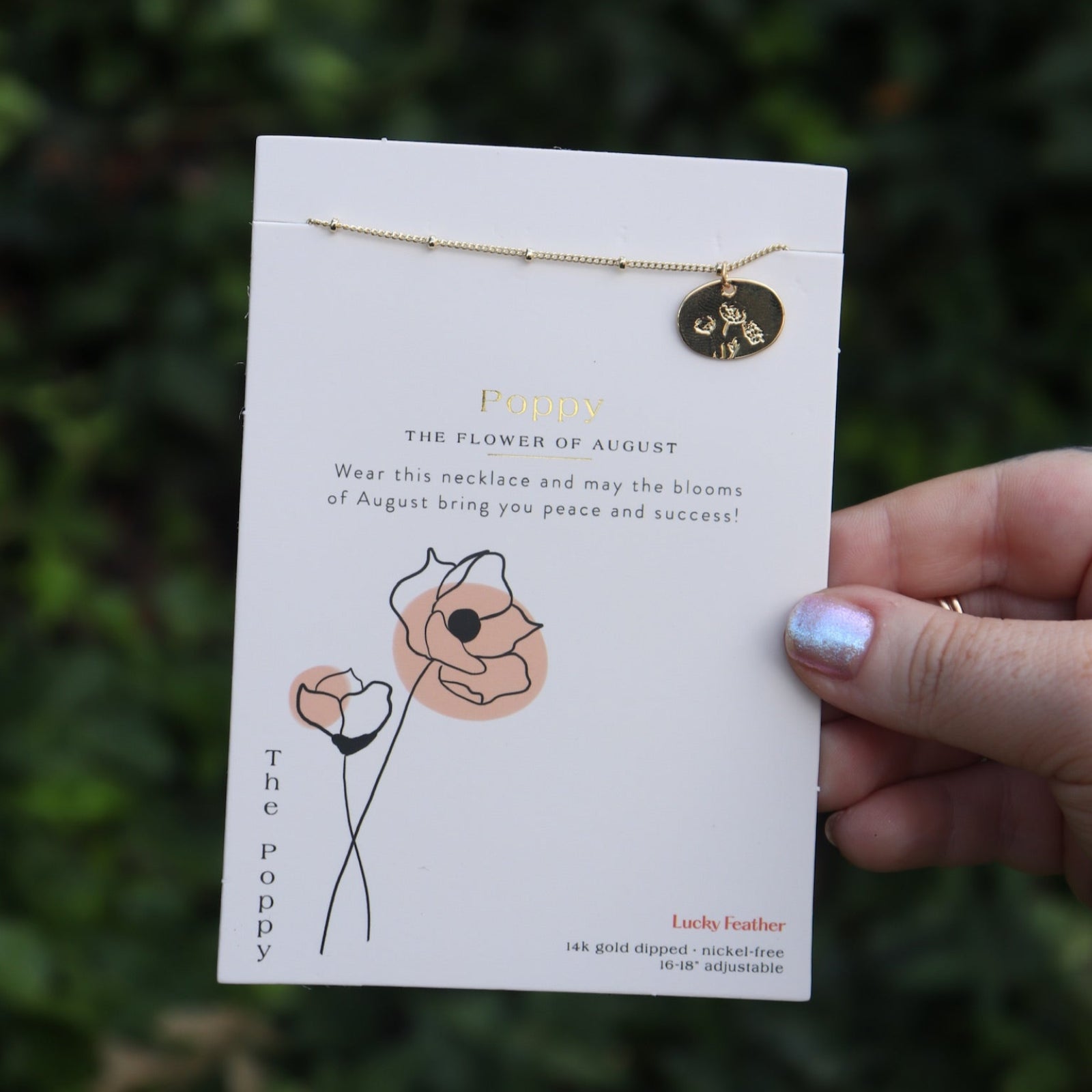 Someone holding the poppy 14k Gold Dipped Nickel-Free Birth Month Flower Necklace in front of greenery.