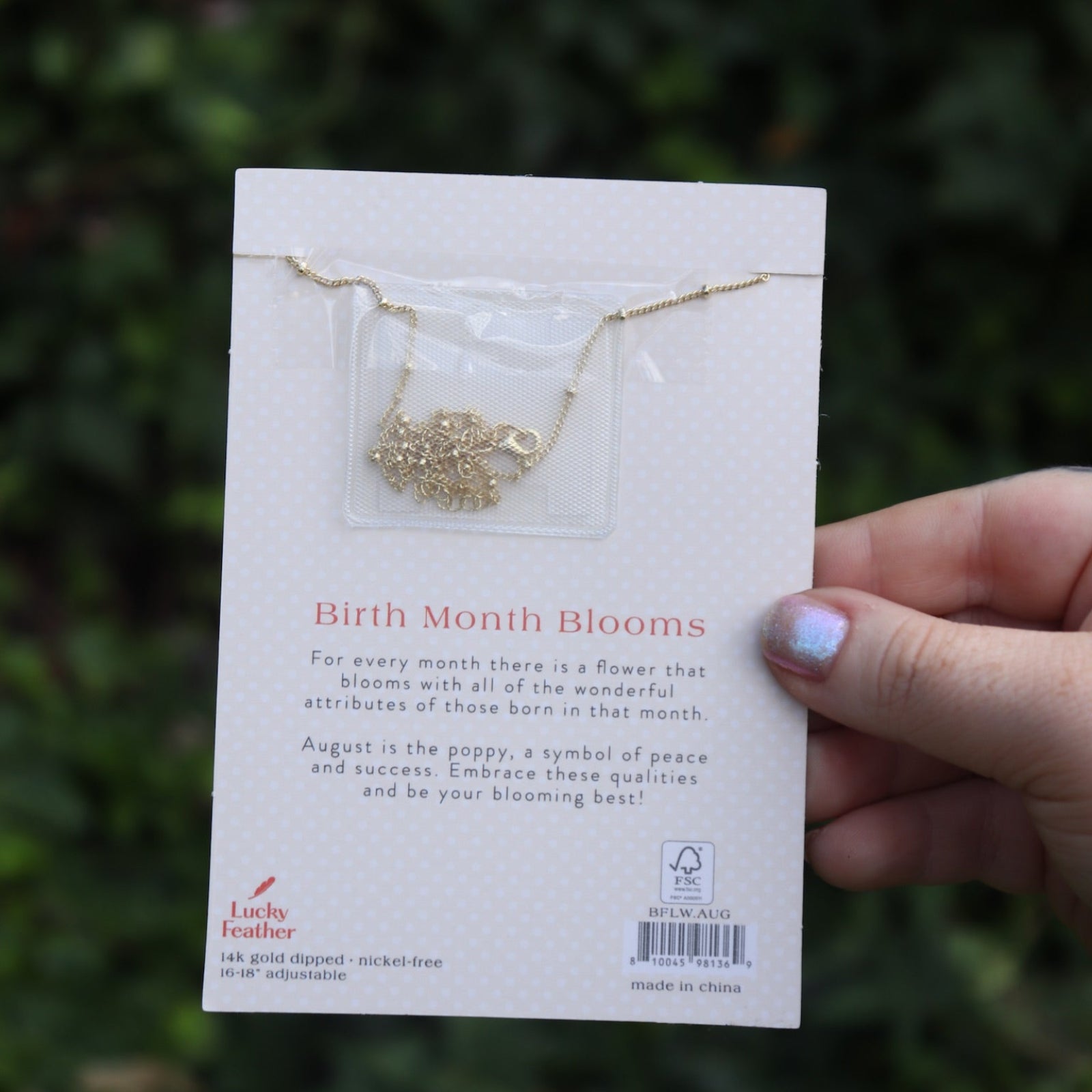 Someone holding the poppy 14k Gold Dipped Nickel-Free Birth Month Flower Necklace in front of greenery showing off the back.