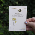 Someone holding the marigold 14k Gold Dipped Nickel-Free Birth Month Flower Necklace in front of greenery.