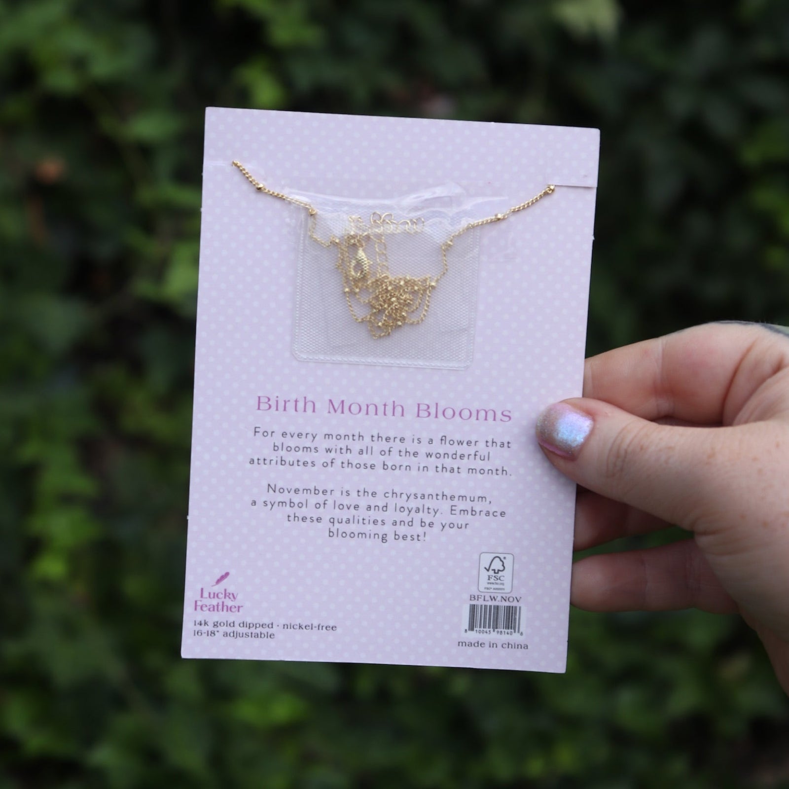 Someone holding the chrysanthemum 14k Gold Dipped Nickel-Free Birth Month Flower Necklace in front of greenery showing off the back.