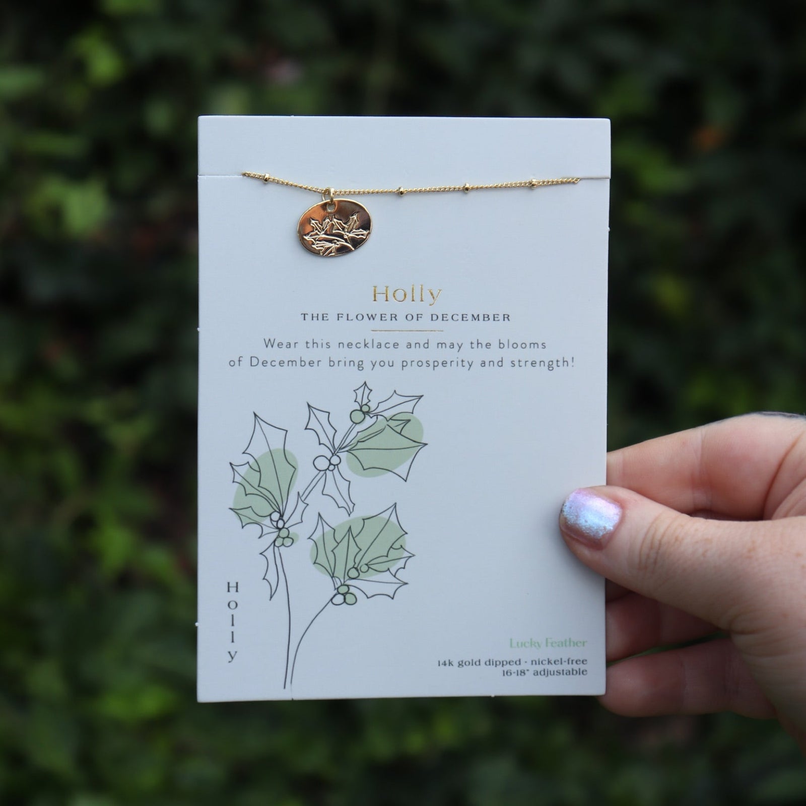 Someone holding the holly 14k Gold Dipped Nickel-Free Birth Month Flower Necklace in front of greenery.