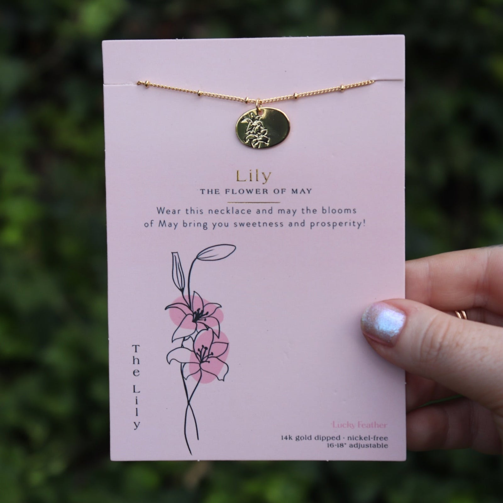 Someone holding the lily 14k Gold Dipped Nickel-Free Birth Month Flower Necklace in front of greenery.