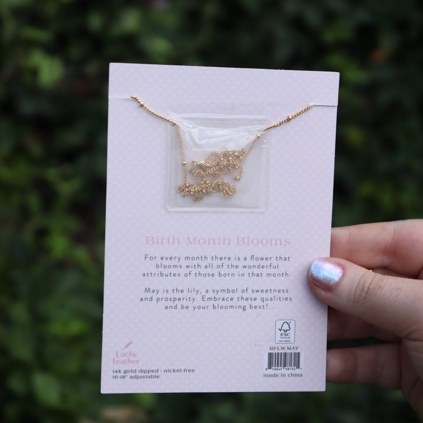 Someone holding the lily 14k Gold Dipped Nickel-Free Birth Month Flower Necklace in front of greenery showing off the back.