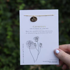 Someone holding the carnation 14k Gold Dipped Nickel-Free Birth Month Flower Necklace in front of greenery.