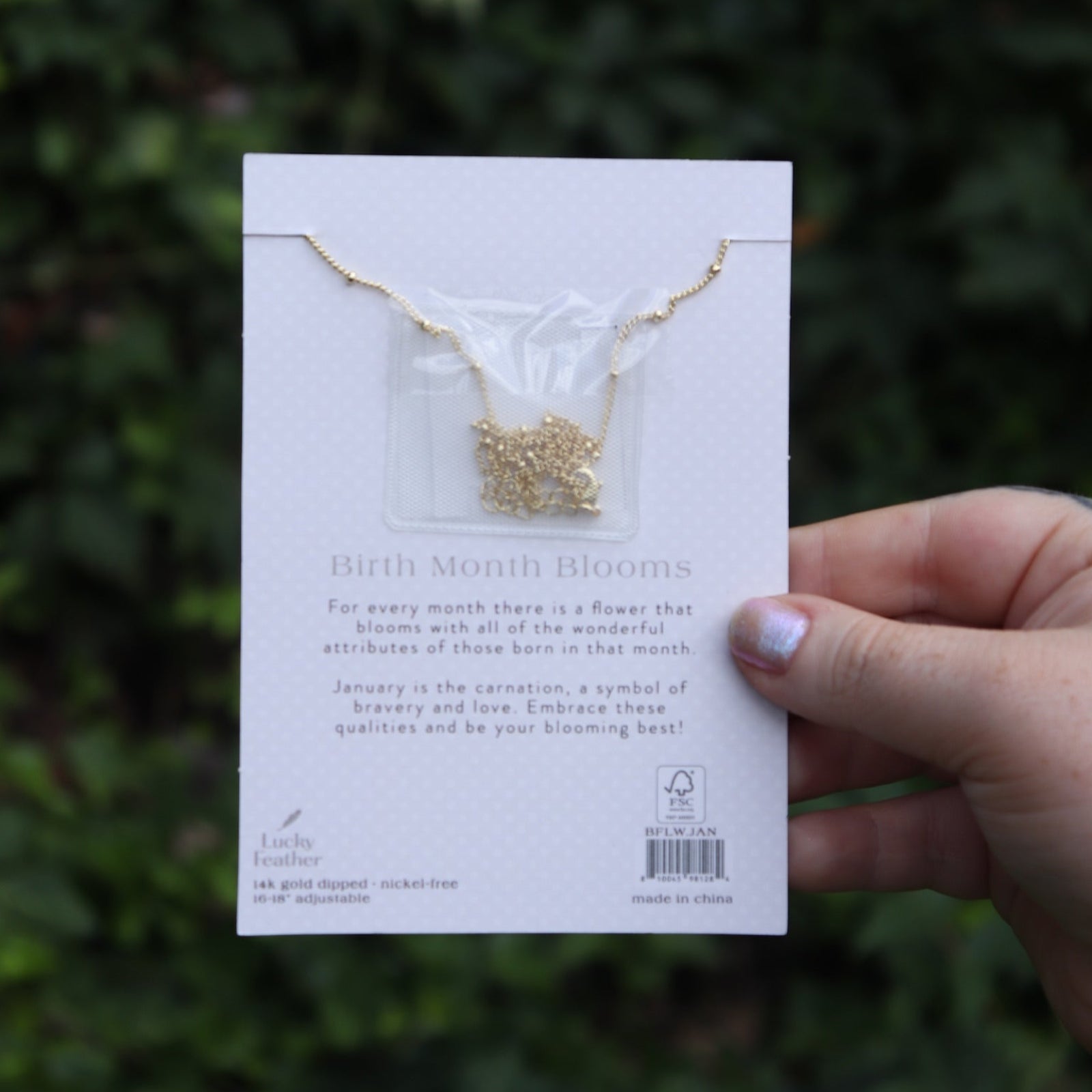 Someone holding the carnation 14k Gold Dipped Nickel-Free Birth Month Flower Necklace in front of greenery showing off the back.
