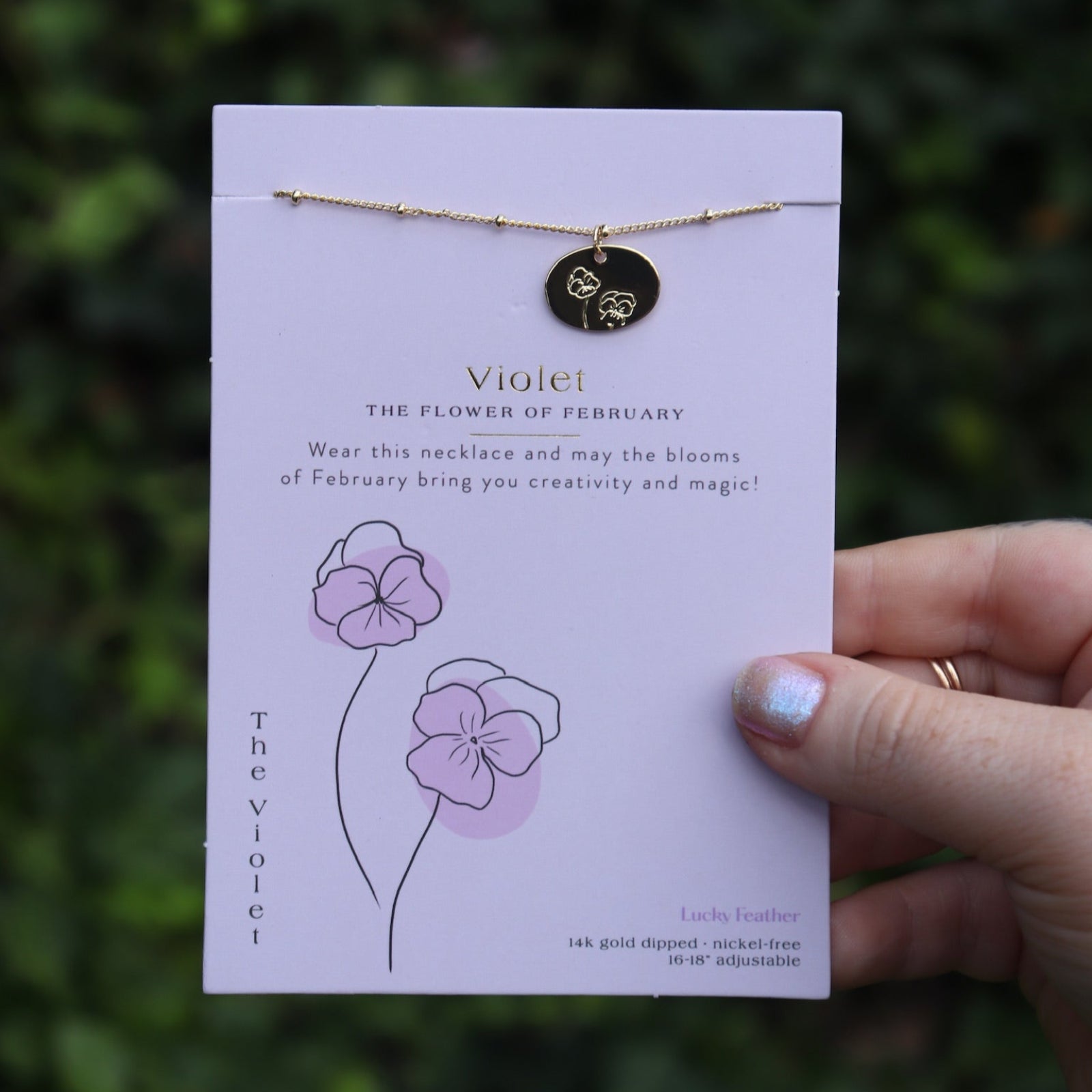 Someone holding the violet 14k Gold Dipped Nickel-Free Birth Month Flower Necklace in front of greenery.